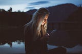 A woman stand outdoors at dusk with her face illuminated by the glow of her cell phone screen