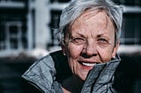 Elderly woman standing outside with a coat on zipped up to the neck.