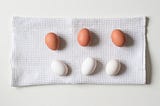 Three brown eggs and three white eggs are arranged in parallel on a white towel cloth on a table.