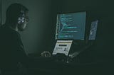 A man wearing headphones in a dimly lit room sitting in front of several computer screens