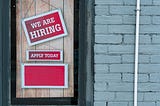 Storefront window with “WE ARE HIRING” and “APPLY TODAY” signs on a red background.