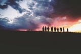 a distant shot of a few people backlit standing on a hill in front of a tumultuous sky