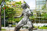 A statue of Bruce Lee, shirtless, in fighting stance, outside a glass building