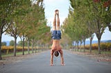 A man performing a handstand in the middle of a tree-lined road, surrounded by autumn foliage. He is shirtless and wearing shorts, with vibrant greenery and a distant landscape visible in the background, creating a serene and balanced composition.