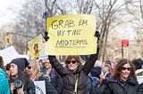 A lady in sunglasses and a black jacket at a protest holding up a sign that says “GRAB EM BY THE MIDTERMS”. There are several others surrounding her and leafless trees in the background indicating it’s wintertime.