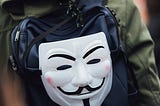 A man with a hooded jacket walking away from the camera. He is wearing a backpack, on which a Guy Fawkes mask hangs, looking directly at the camera.