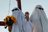 A couple holding hands with flowers and putting on white blankets as ghost costumes with sunglasses on