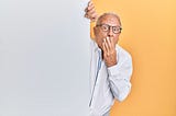 An old man with glasses peeking from behind a wall with his right hand on his mouth and eyes wide with shock