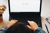 Hands typing on a black laptop displaying a Google screen