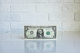 A $1 bill sits upright on a white counter top in front of a white brick wall