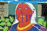 A colourful wall mural of an elder’s face. They have long white hair and markings on their left cheek and from nose to chin in purple.