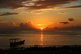 The sun peeking up on the horizon, over an ocean with a humble boat anchored at the rocky shore.