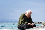 Old man sitting on a beach and looking down, appearing to be sad.