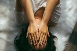 An aerial view of a woman in a white tee shirt laying on a crumpled white sheeted bed, her hands covering her face.