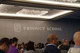 Conference room filled with people sitting down for a session with the Product School visible on a grey wall in the distance.