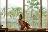 Girl sits by a window looking at a hot, tropical view.