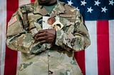 photo of person in camoflauge uniform with american flag in background, holding photos of a baby and a woman, posed on mickey markoff 2024 article on day of the deployed