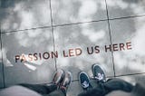 sidewalk with “Passion Led Us Here” on it, two pairs of feet point to the letter.