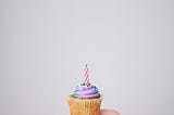 A hand holding a cupcake with a candle on it.