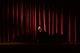 A person sitting behind the closed red curtain on a stage, looking to his right.