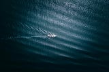 A small sailboat moves alone through dark blue ocean waters, leaving ripples behind it.