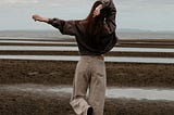 A woman stands on one foot, one arm extended, the other crooked behind her head. She faces away from us, towards streaks of land and water. She wears a dark sweater and beige long pants.