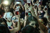 Party scene with a group of people raising glasses filled with red alcohol in the air. In the background, guests are filming on their phones and in the foreground, you can see part of a lady’s face, but she looks distressed.