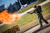 A man fires a flamethrower at an elementary school track field. He imagines what it would be like to be young again instead of committing crimes on public property. He probably created an NFT project that didn’t add any value to his community and is now doomed to commit public displays of insanity and poor weapons safety.