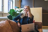 A blonde woman sitting on a brown leather sofa. She wears a dark blue suit, is talking on her mobile phone and has an Apple computer on her lap. She is smiling and looking efficient.