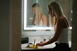 Young blond woman alone in a bathroom, standing in front of a mirror.