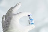 A close-up of a person’s hand wearing a white latex glove, gently holding a small glass vial between thumb and forefinger against a soft, light blue-gray background. The vial has a blue-and-silver cap and a white label reading “SARS-CoV-2 mRNA Vaccine COVID-19,” with “EXP 2021–06–03” printed vertically along the side.