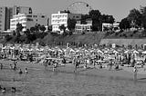 A black and white view of a beach, taken from the sea. The sand is crowded with sunshades; low-rise apartment blocks or hotels stand in the background.