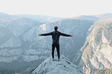 A person standing with arms out on a high hill looking out at mountains beyond.