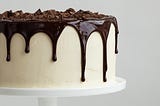 Photo of a vanilla cake on a cake platter with chocolate ganache dripping down the sides and shaved chocolate on top.