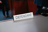 A plain white sign with black lettering sits on a table. It says Defendant.