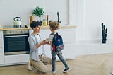 A mother standing in the kitchen with her son, wishing him luck for his first day of school.