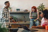 A smiling black man in a white checked shirt faces a red haired woman and a black woman in a kitchen. The red haired woman is sitting on a kitchen counter and the black woman is sitting at a counter. Both women are smiling.
