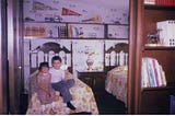 A little girl of 3 is sitting on the edge of a bed. Her older brother is hugging her and holding a paper airplane.