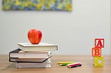 teacher’s desk with a pile of books, an apple, scattered colored pencils, and A B C wooden blocks