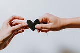 One hand reaches out from the left side of this image to recieve a black cutout heart being handed to them by somone else on the right. The background is a plain white.