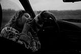 A greyscale photo of a woman smoking a cigarette in a car as it rains.