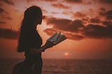 A woman holds an open book, staring at a sunset over the ocean.