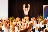 Scores hands raised in the air and a speaker with a microphone in the distance, on stage, hands raised and addressing the room