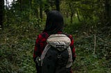 Person with long black hair standing in a forest. Wearing a hiking backpack, and red flannel shirt.