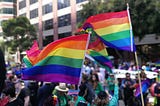 A crowd of people on a street, none clearly visible, with three rainbow flags in the foreground.