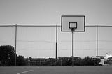 A black-and-white image of an empty outdoor basketball hoop and court, with a chain-link fence in the background.