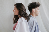 Man and woman standing back to back with somber looks on their faces.