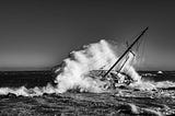 Black and white photo of an abandoned sailboat with a wave crashing over it.
