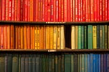 A shelf of books arranged by colour from orange to red, yellow, green, then blue.