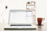 A laptop and a mug of tea on top of a study table in a brightly lit room.